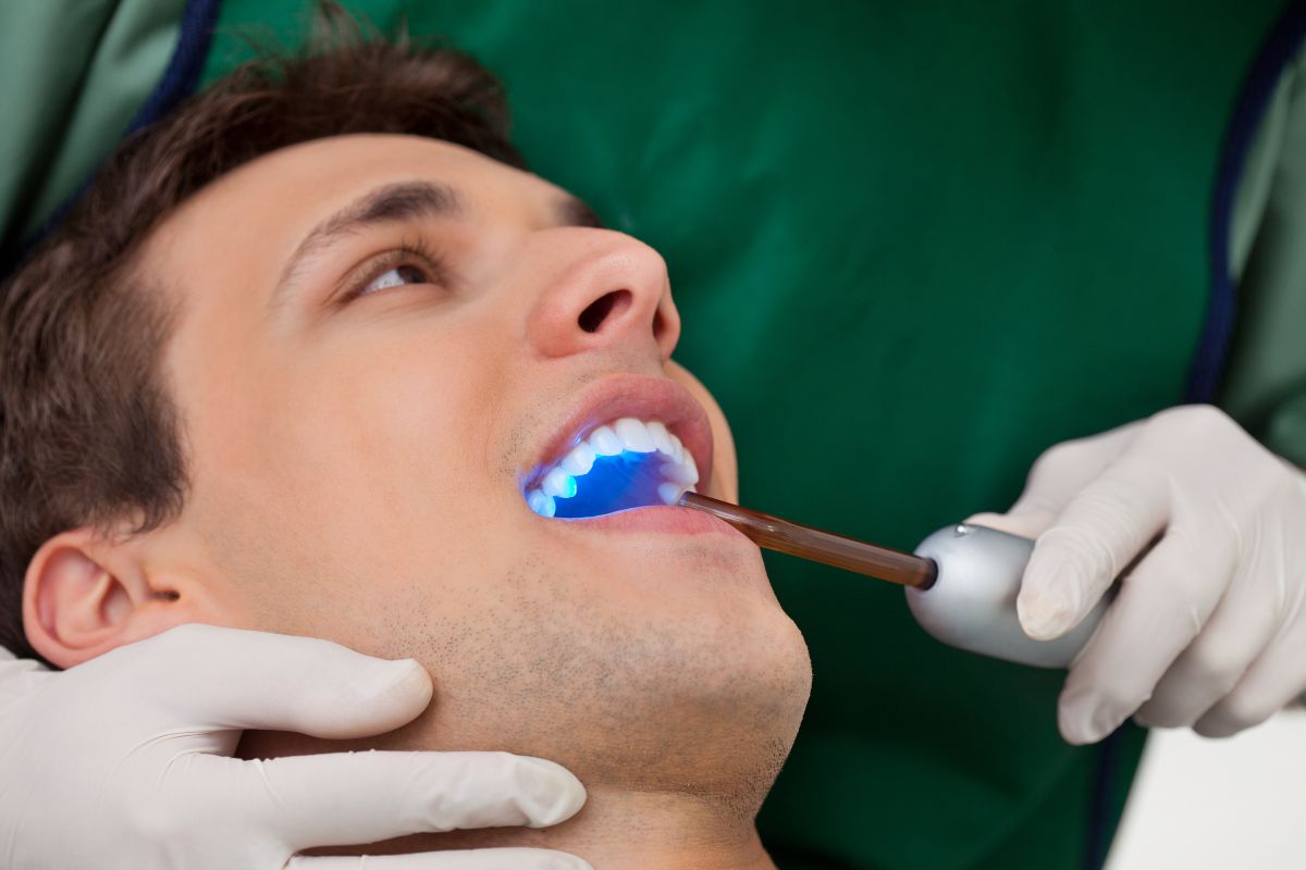 Dentist performing teeth whitening procedure on male patient using blue light technology in dental office.