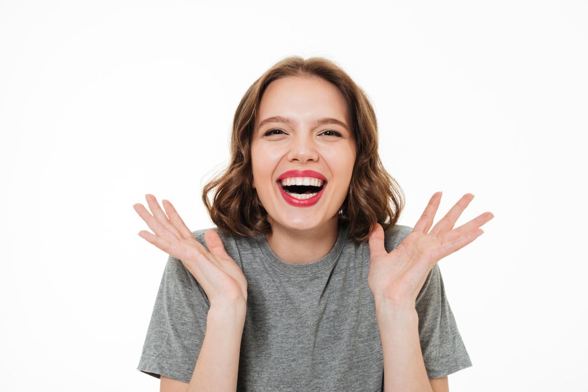 Excited woman in gray shirt with hands raised, smiling against white background
