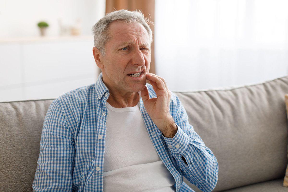 How to Handle a Dental Emergency Mature man sitting on couch, thoughtfully touching face, wearing casual blue checkered shirt.